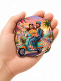 Fridge Magnet of a couple on a scooter with a tropical background held in a hand.
